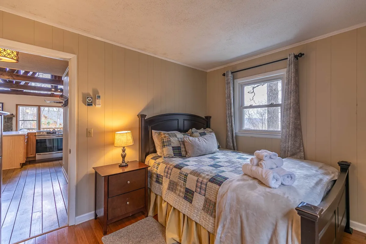 A tranquil bedroom with a bed featuring a quilted cover, a nightstand with a lamp, and a window that floods the room with natural light. The mood is restful, ideal for quiet mornings or peaceful nights.