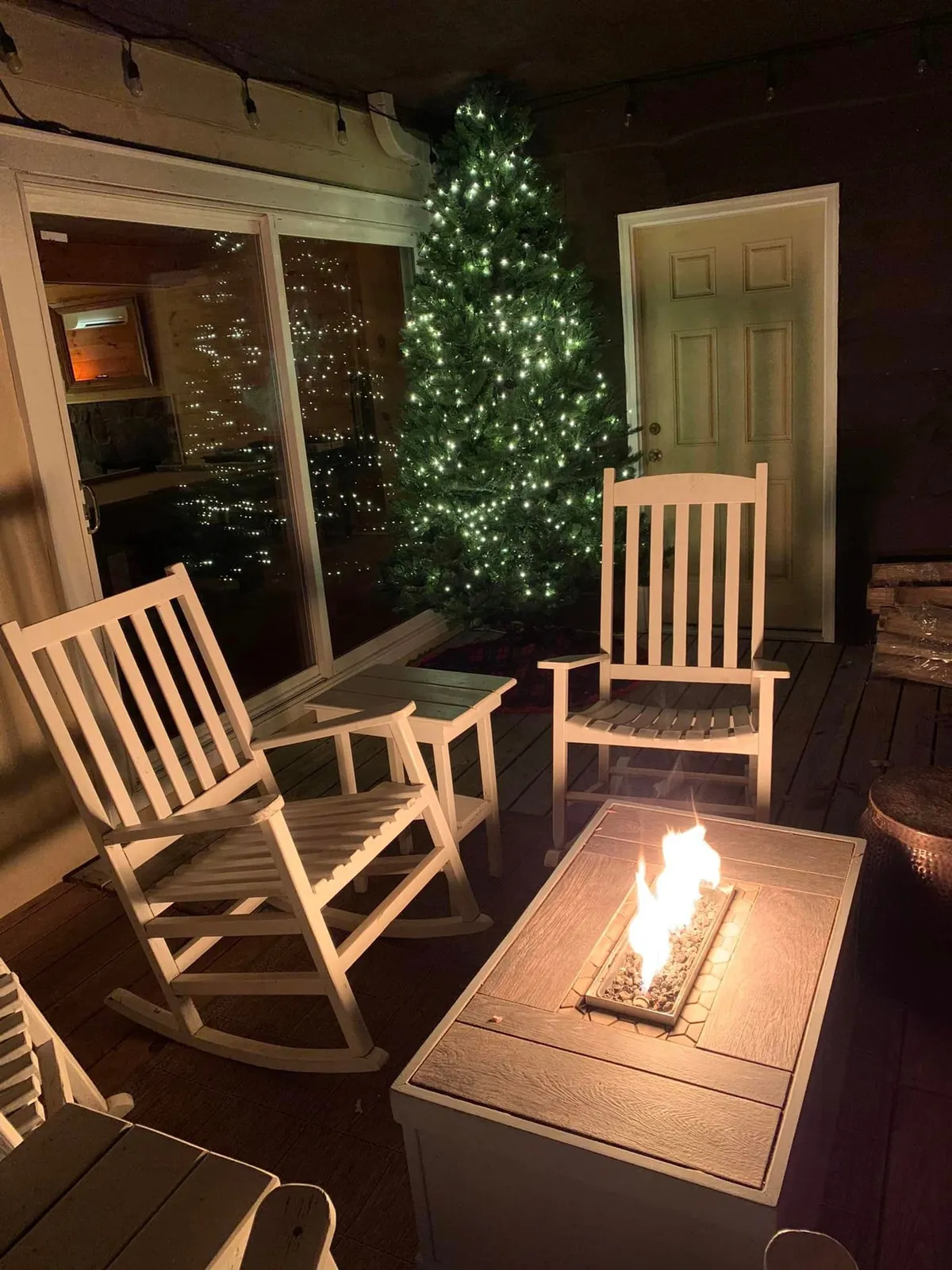A cozy outdoor fire pit area is surrounded by comfortable rocking chairs, set beneath the trees and adjacent to a glowing Christmas tree. The warm, inviting scene suggests storytelling, stargazing, and close-knit group moments around the fire.