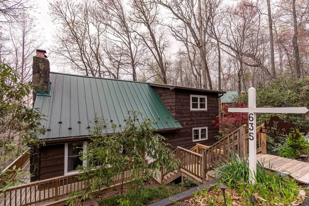 A charming wooden cabin with a green metal roof sits nestled among the trees, its wraparound deck and inviting signpost creating a welcoming first impression. The lush greenery and open yard suggest ample space for outdoor gatherings and family relaxation.