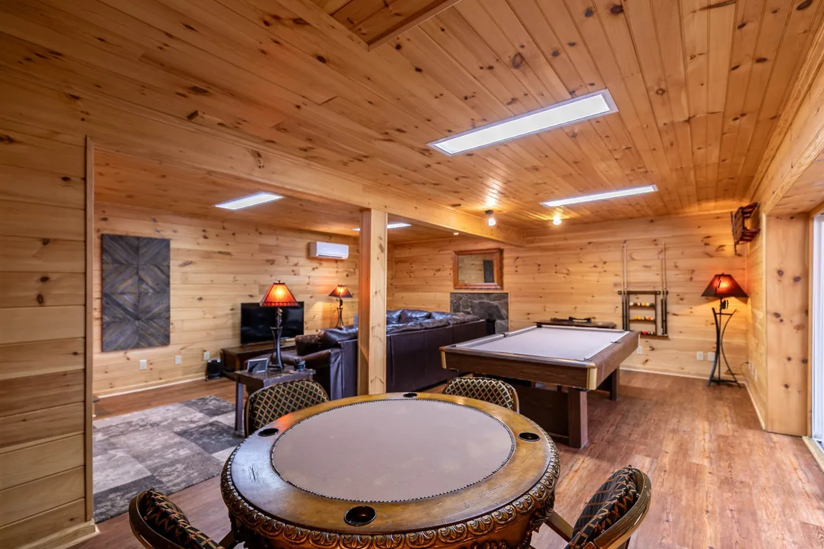This image presents a vibrant lower-level game room featuring a pool table, a comfortable circular table with chairs for board games, a cozy sofa area with a TV, and rustic wooden walls that add warmth to the space. The layout showcases distinct zones for both gaming and relaxing, with soft lighting that creates a welcoming ambiance perfect for group fun.