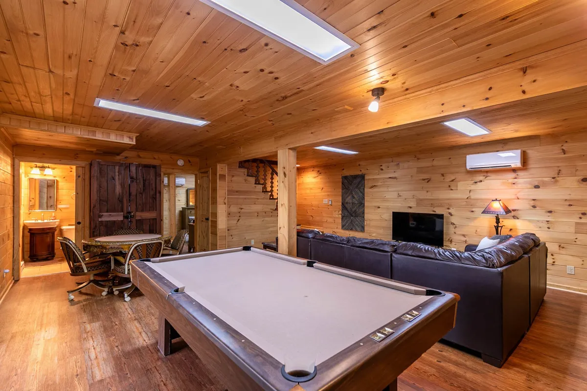 The scene captures a relaxed lower-level space with a pool table in the foreground and a leather sofa set up for comfortable socializing or movie watching. Wooden decor elements and a dining table add to the overall communal feel, while the arrangement encourages both spirited gameplay and cozy gatherings.