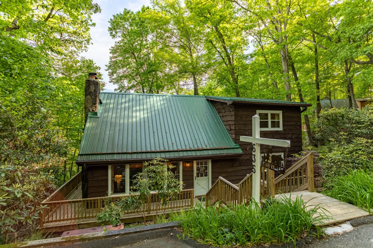 An exterior shot of a charming wooden house with a green roof and a welcoming front porch. The home is nestled among trees, giving it a serene, inviting feel. There’s a signpost beside the porch, and the setting suggests easy access to nature.