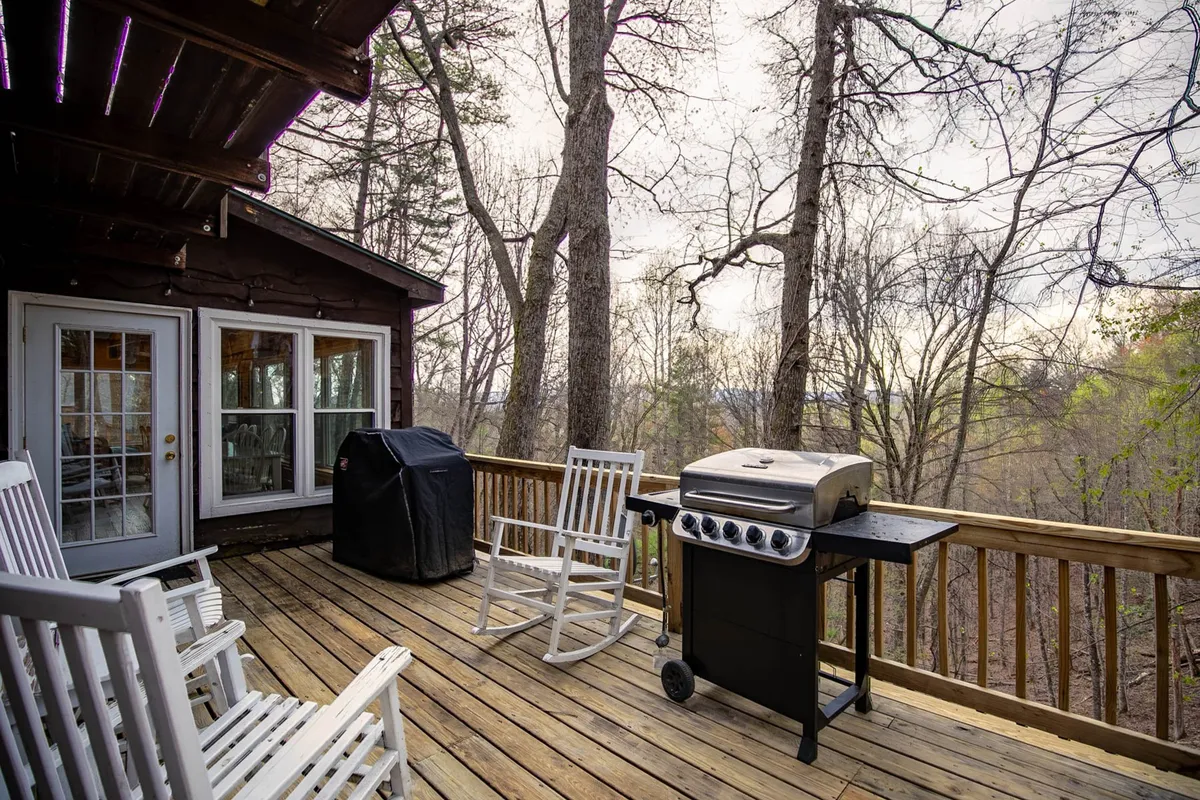 A wooden deck surrounded by trees features a grill and classic rocking chairs, creating the ideal setting for outdoor meals, barbecues, and relaxed socializing with loved ones. The tranquil natural backdrop enhances the feeling of a mountain getaway.