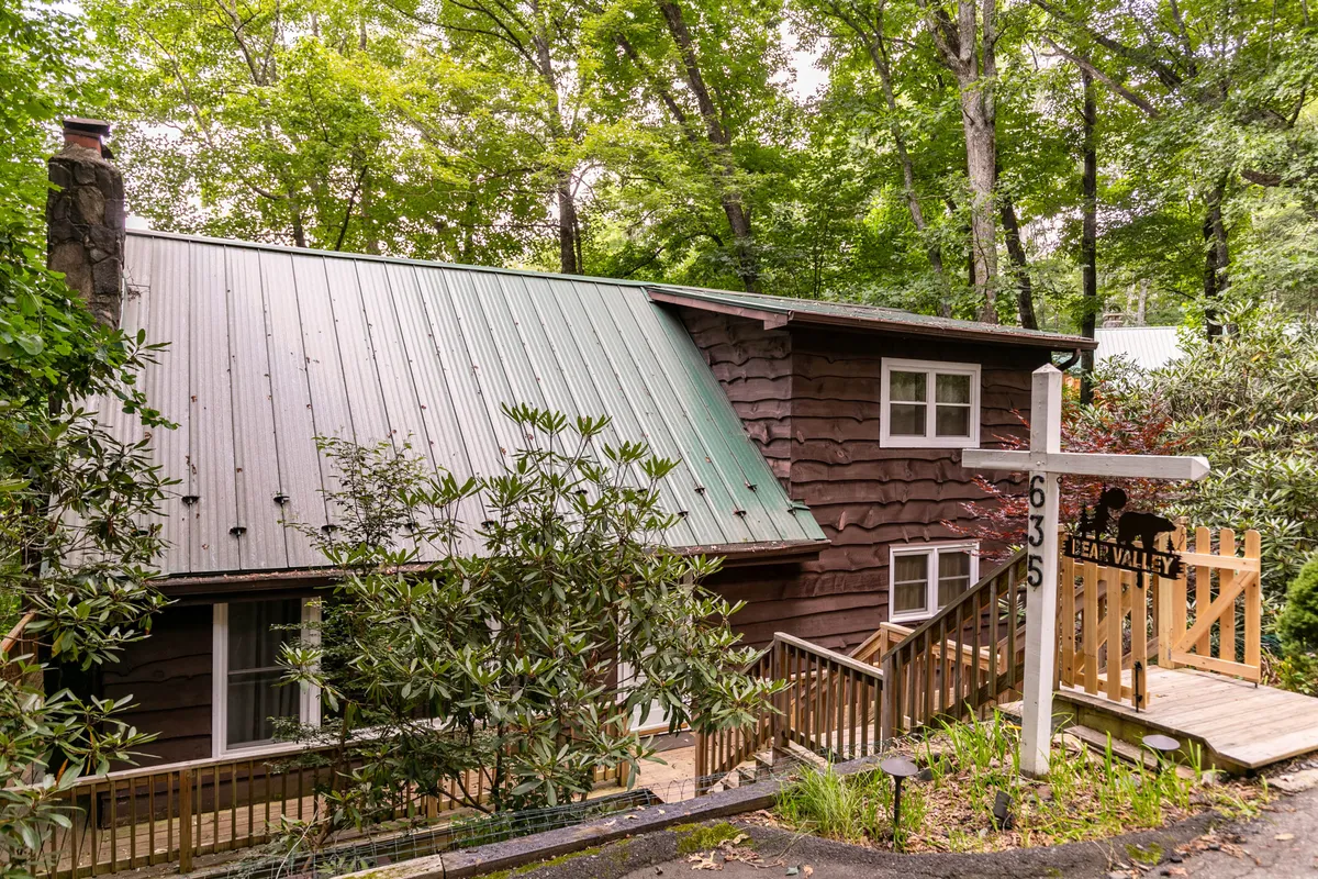 A view of the home's wooden exterior and green metal roof, with a welcoming pathway leading toward the entrance. The setting is lush and surrounded by greenery, suggesting a tranquil spot for summer relaxation and outdoor adventures.