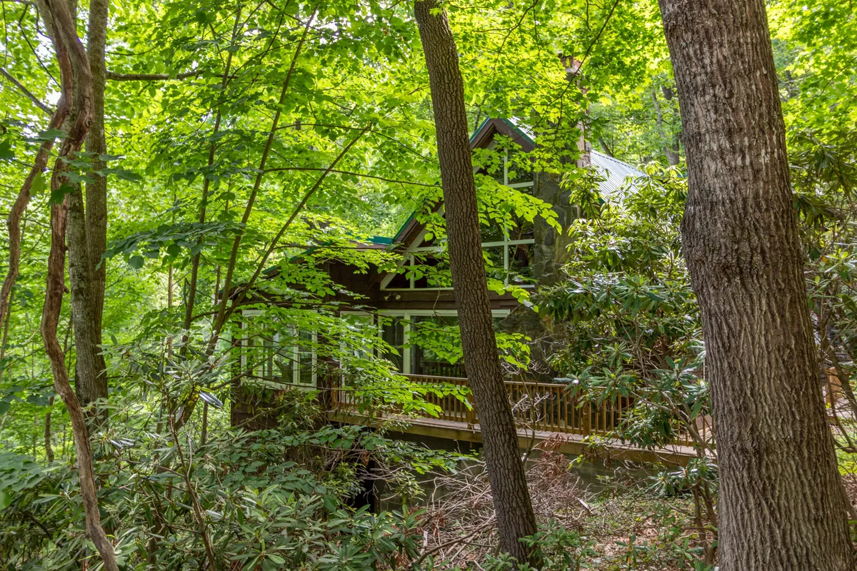 The image shows the house nestled among lush trees with a spacious deck extending out, surrounded by serene mountain scenery. The area evokes peace and privacy, inviting guests to relax and take in the natural surroundings.