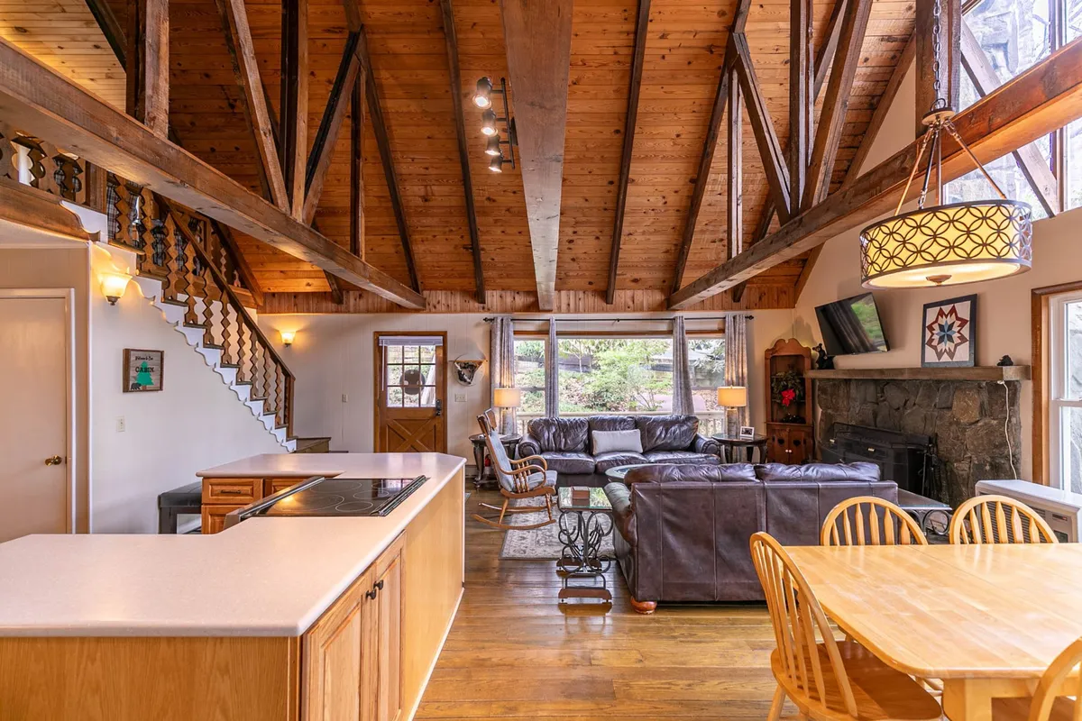 The great room's dramatic vaulted wooden ceiling is accented by chunky exposed beams. A large leather sectional sofa wraps around the stone fireplace, while a wooden dining table and open kitchen are visible nearby, all bathed in natural light from oversized windows.