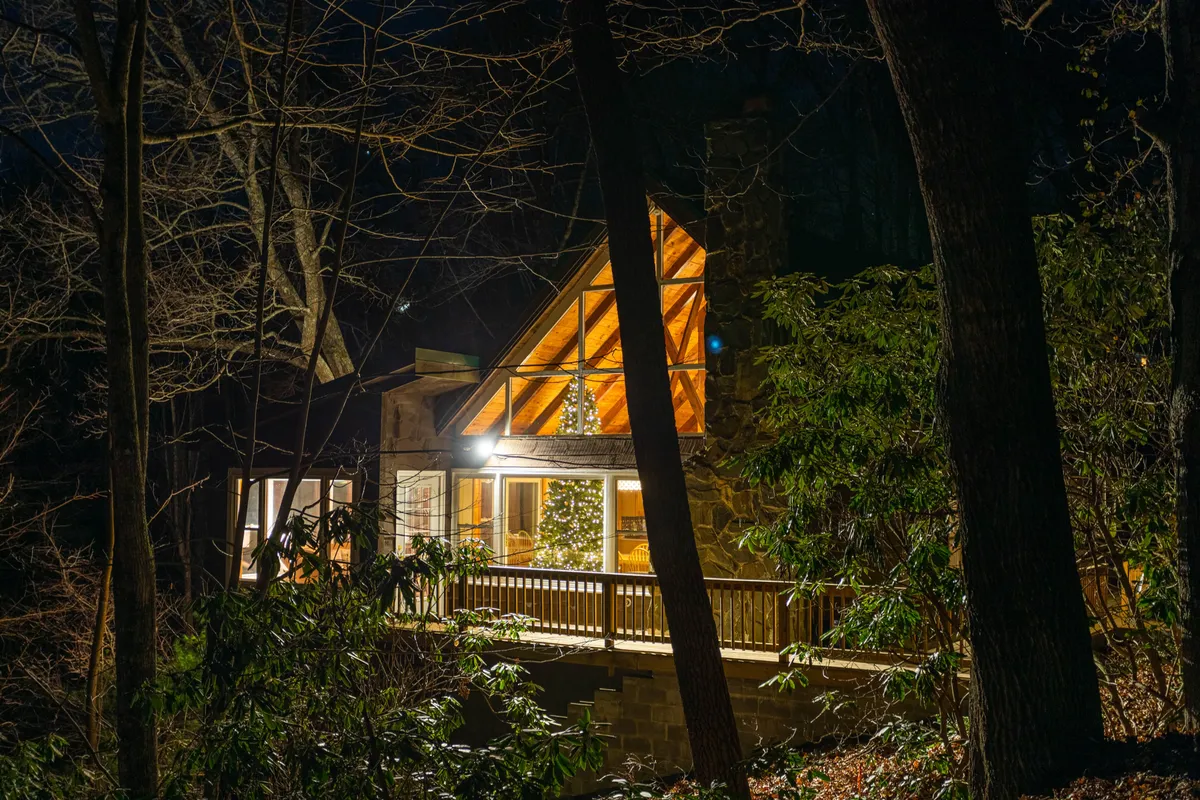 A warmly lit house framed by trees at dusk, with the soft glow of lights visible through the windows and a decorated Christmas tree displayed inside. The wooden deck and the surrounding natural landscape evoke a sense of seasonal coziness and outdoor connection.