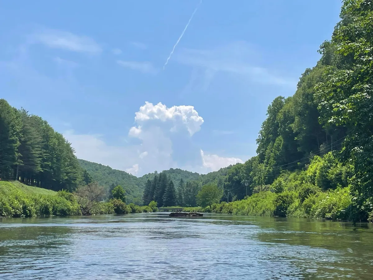 A sweeping view of the nearby river, with clouds reflected on the water, green hills, and trees along the shore. The tranquil scene suggests outdoor activities like hiking and picnicking, and evokes the peaceful mountain air and riverside access described in the section.