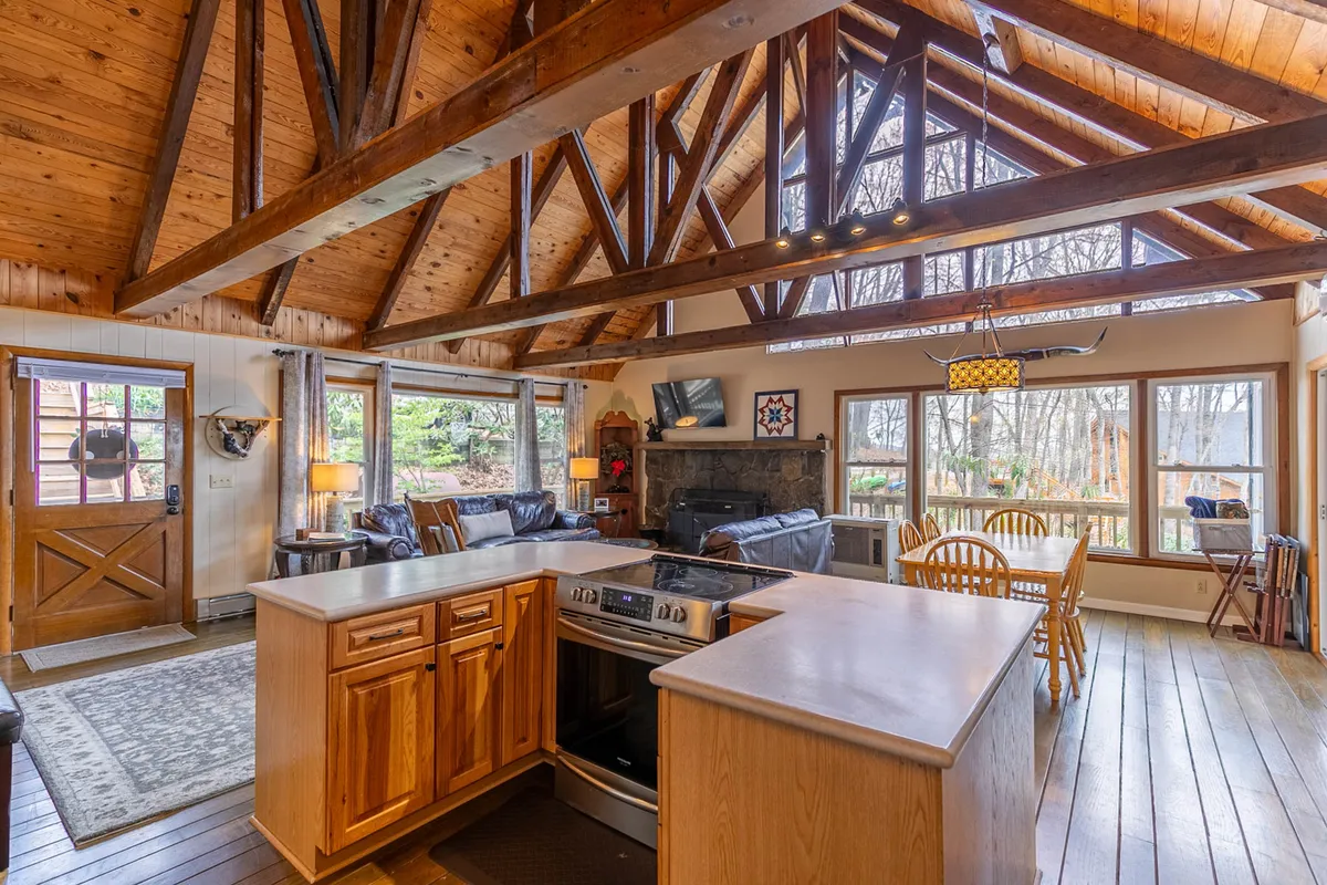This expansive great room showcases a soaring vaulted wooden ceiling with prominent exposed beams, filling the space with natural light from large windows. The inviting stone fireplace anchors the living area, while a modern kitchen island and a spacious dining area are seamlessly integrated for easy gatherings. The room exudes warmth, making it ideal for family meals, cooking together, or relaxing by the fire.