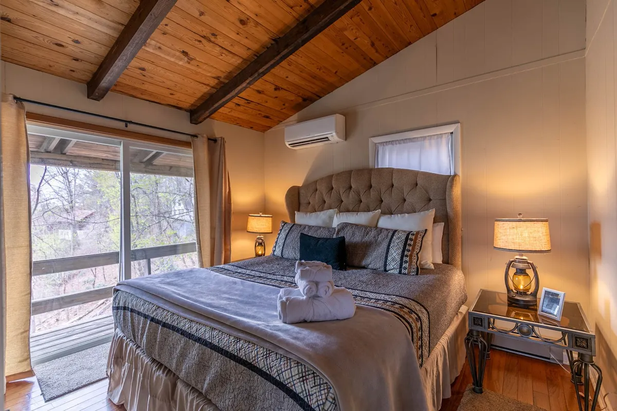 A serene bedroom featuring a king-size bed, large windows, and soft decorative pillows. Side tables with lamps add to the ambiance, while the view out to nature hints at a tranquil retreat.